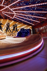 A brightly lit amusement ride featuring spinning motion. The scene is adorned with numerous lightbulbs arranged in patterns, creating vibrant light trails. Silhouettes of dancers are painted against a yellow backdrop, while the ceiling is covered in star patterns. The ride structure is red, with blue and white colors prevalent in the lighting.