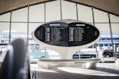 An airport terminal features a large, retro departure and arrival board displaying various flight destinations and times. The architectural design is futuristic, with sweeping curves and a minimalist aesthetic. Automobiles and a vintage Volkswagen van are visible through the large windows, adding to the scene.