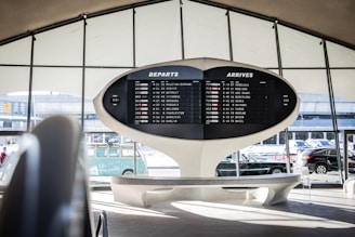 An airport terminal features a large, retro departure and arrival board displaying various flight destinations and times. The architectural design is futuristic, with sweeping curves and a minimalist aesthetic. Automobiles and a vintage Volkswagen van are visible through the large windows, adding to the scene.