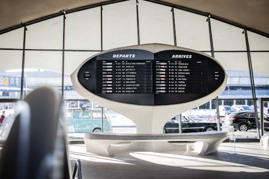 An airport terminal features a large, retro departure and arrival board displaying various flight destinations and times. The architectural design is futuristic, with sweeping curves and a minimalist aesthetic. Automobiles and a vintage Volkswagen van are visible through the large windows, adding to the scene.
