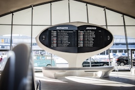 An airport terminal features a large, retro departure and arrival board displaying various flight destinations and times. The architectural design is futuristic, with sweeping curves and a minimalist aesthetic. Automobiles and a vintage Volkswagen van are visible through the large windows, adding to the scene.