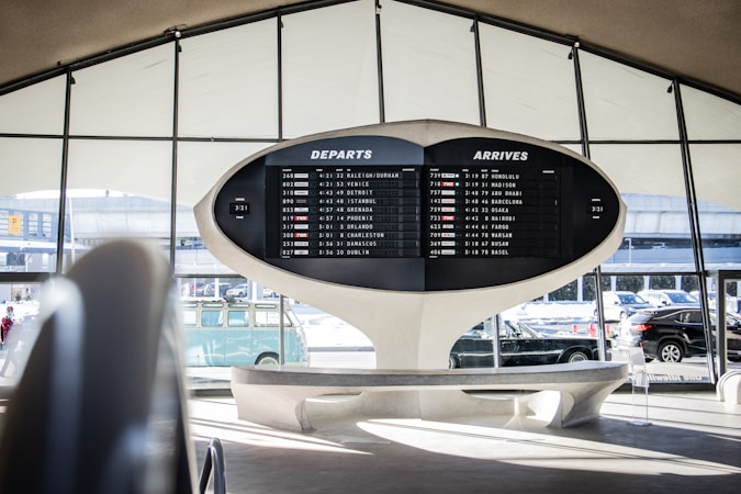 An airport terminal features a large, retro departure and arrival board displaying various flight destinations and times. The architectural design is futuristic, with sweeping curves and a minimalist aesthetic. Automobiles and a vintage Volkswagen van are visible through the large windows, adding to the scene.