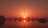 A stunning sunset behind a futuristic cityscape with tall, spire-like structures reflecting on a calm body of water. The warm orange and red hues create a serene and awe-inspiring atmosphere.