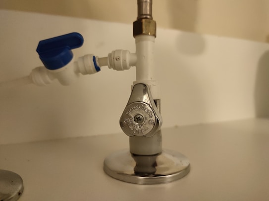 A close-up view of plumbing components including a shut-off valve with a metallic handle labeled 'OFF' on a pipe, adjacent to a blue-handled plastic valve.