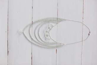 A delicate silver necklace with intricate beadwork is displayed on a rustic white wooden surface. The necklace features multiple strands that converge into a series of small, translucent beads.