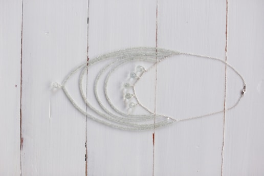 A delicate silver necklace with intricate beadwork is displayed on a rustic white wooden surface. The necklace features multiple strands that converge into a series of small, translucent beads.
