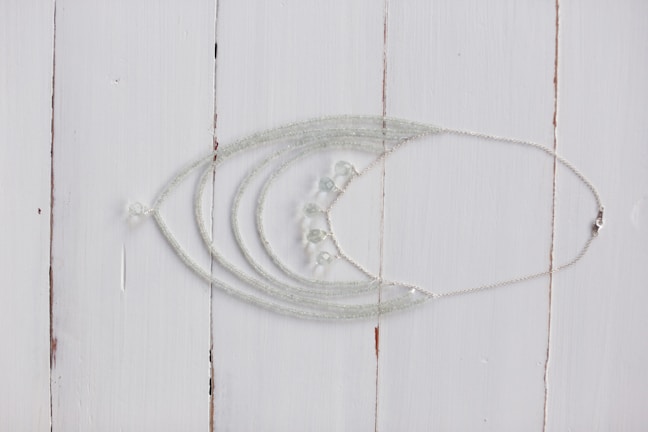 A delicate silver necklace with intricate beadwork is displayed on a rustic white wooden surface. The necklace features multiple strands that converge into a series of small, translucent beads.