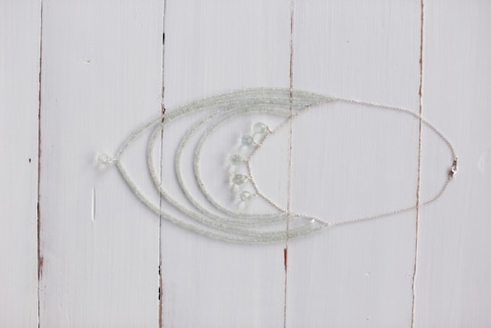 A delicate silver necklace with intricate beadwork is displayed on a rustic white wooden surface. The necklace features multiple strands that converge into a series of small, translucent beads.