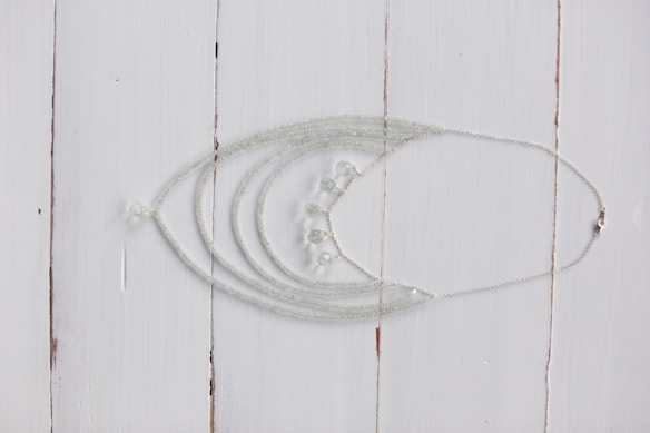 A delicate silver necklace with intricate beadwork is displayed on a rustic white wooden surface. The necklace features multiple strands that converge into a series of small, translucent beads.