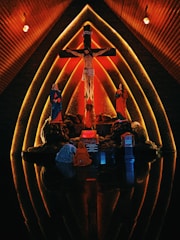 The image features a religious scene set against a dramatic, warmly lit background. At the center is a depiction of Jesus on the cross, flanked by two figures in red and blue robes. The setting appears to be inside a church with wooden, arched walls that create a halo-like effect around the figures, emphasizing the sacred nature of the scene.
