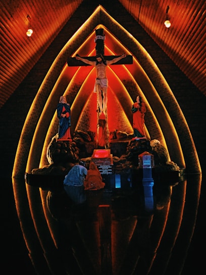The image features a religious scene set against a dramatic, warmly lit background. At the center is a depiction of Jesus on the cross, flanked by two figures in red and blue robes. The setting appears to be inside a church with wooden, arched walls that create a halo-like effect around the figures, emphasizing the sacred nature of the scene.