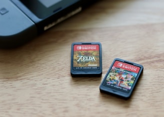 Two Nintendo Switch game cartridges are placed on a wooden surface near a corner of a Nintendo Switch console. The cartridge on the left features the game 'The Legend of Zelda: Breath of the Wild' while the one on the right displays 'Mario Kart 8 Deluxe'. The surface is light-colored, providing contrast to the dark cartridges.