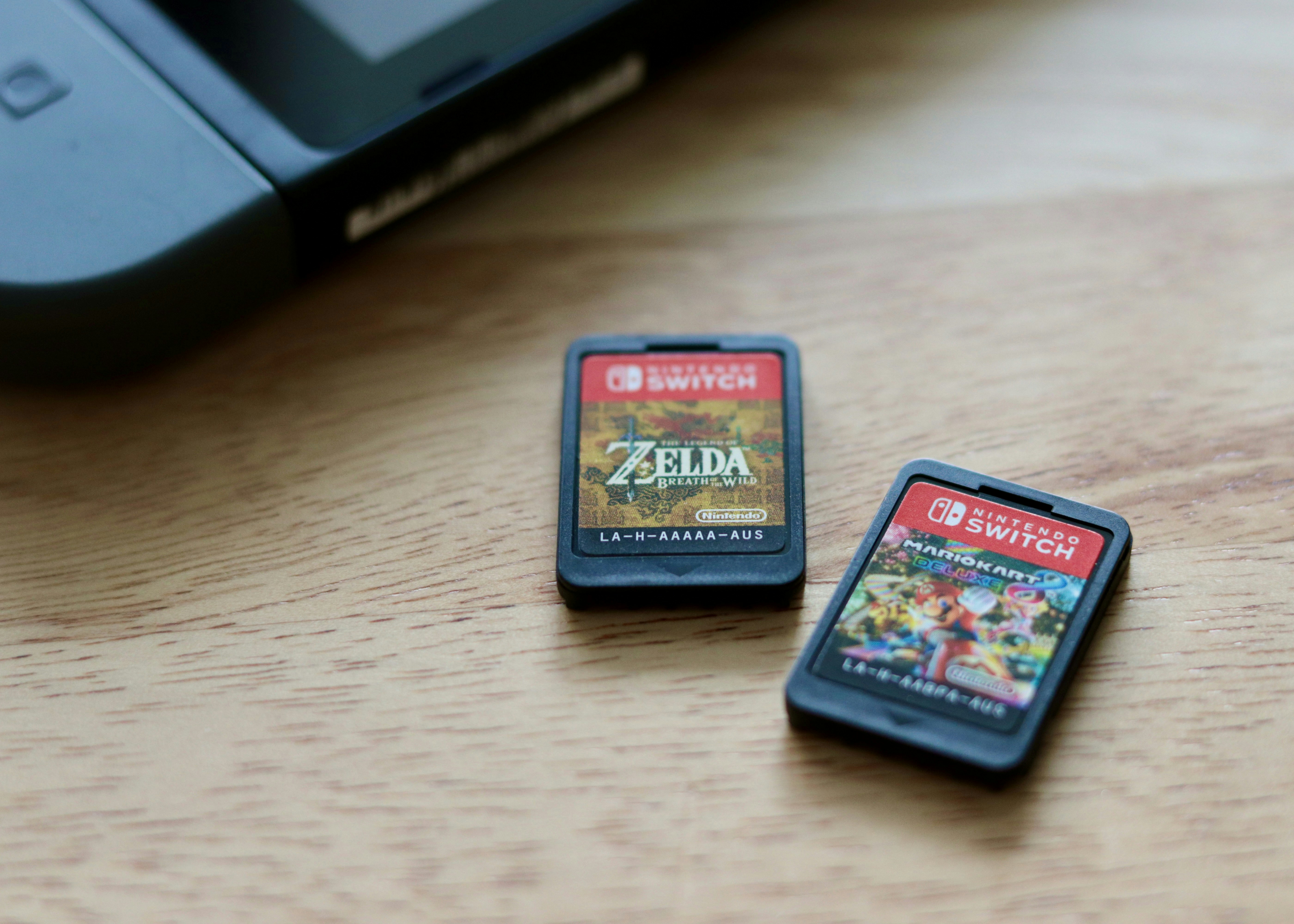 Two video game cartridges for the Nintendo Switch laying on a wooden table. The one at the front is for Mario Kart 8; the one on the left is for The Legend of Zelda: Breath of the Wild. Also in the background is the corner of a black Nintendo Switch console in its handheld mode, with both controllers locked onto the body of the device.