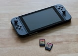 A portable gaming console is placed on a wooden surface, accompanied by two small game cartridges. The console features a screen in the center with controllers on either side, while the cartridges have colorful labels depicting game titles.