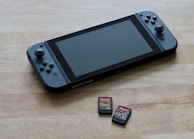 A portable gaming console is placed on a wooden surface, accompanied by two small game cartridges. The console features a screen in the center with controllers on either side, while the cartridges have colorful labels depicting game titles.
