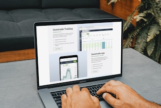 A person is using a MacBook Pro, viewing financial trading information from Questrade. The screen displays detailed trading data and an app interface, emphasizing connectivity and accessibility. The image features a modern setting with a couch in the background and some houseplants.