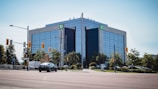 A modern office building with reflective glass windows stands prominently at the intersection. The building is surrounded by greenery and traffic lights, with a few vehicles on the road. A 'TD' sign is visible at the top corner of the building.