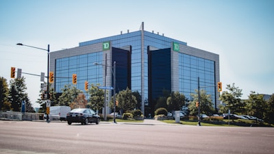 A modern office building with reflective glass windows stands prominently at the intersection. The building is surrounded by greenery and traffic lights, with a few vehicles on the road. A 'TD' sign is visible at the top corner of the building.