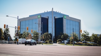 A modern office building with reflective glass windows stands prominently at the intersection. The building is surrounded by greenery and traffic lights, with a few vehicles on the road. A 'TD' sign is visible at the top corner of the building.