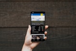 A hand holding a smartphone displaying a financial website, with information including market watch and stocks data visible on the screen. The background is a textured wooden surface.