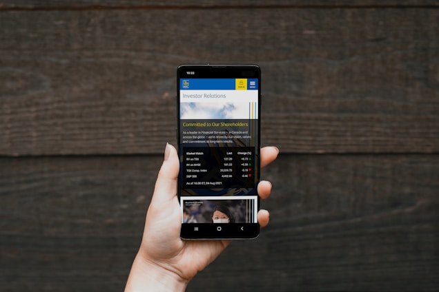 A hand holding a smartphone displaying a financial website, with information including market watch and stocks data visible on the screen. The background is a textured wooden surface.