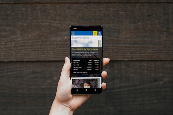 A hand holding a smartphone displaying a financial website, with information including market watch and stocks data visible on the screen. The background is a textured wooden surface.