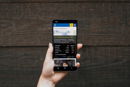 A hand holding a smartphone displaying a financial website, with information including market watch and stocks data visible on the screen. The background is a textured wooden surface.