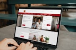 A person is using a MacBook Pro laptop displaying a bank's website. The screen shows sections for logging in, credit card offers, and various banking services. The user interface is predominantly white with red accents, and the background consists of a wooden table and seating.