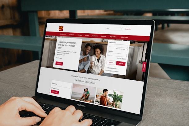 A person is using a MacBook Pro laptop displaying a bank's website. The screen shows sections for logging in, credit card offers, and various banking services. The user interface is predominantly white with red accents, and the background consists of a wooden table and seating.