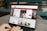 A person is using a MacBook Pro laptop displaying a bank's website. The screen shows sections for logging in, credit card offers, and various banking services. The user interface is predominantly white with red accents, and the background consists of a wooden table and seating.
