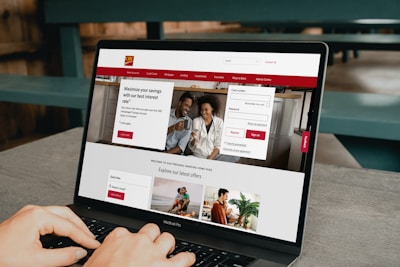 A person is using a MacBook Pro laptop displaying a bank's website. The screen shows sections for logging in, credit card offers, and various banking services. The user interface is predominantly white with red accents, and the background consists of a wooden table and seating.