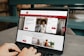 A person is using a MacBook Pro laptop displaying a bank's website. The screen shows sections for logging in, credit card offers, and various banking services. The user interface is predominantly white with red accents, and the background consists of a wooden table and seating.