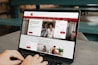 A person is using a MacBook Pro laptop displaying a bank's website. The screen shows sections for logging in, credit card offers, and various banking services. The user interface is predominantly white with red accents, and the background consists of a wooden table and seating.