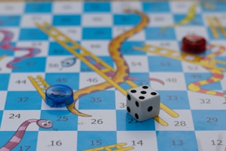 A board game features a grid with alternating blue and white squares, decorated with illustrated snakes and ladders. A six-sided die and circular game pieces in red and blue are placed on the board. The squares display numbers, adding a structured element to the colorful, playful design.