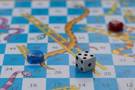 A board game features a grid with alternating blue and white squares, decorated with illustrated snakes and ladders. A six-sided die and circular game pieces in red and blue are placed on the board. The squares display numbers, adding a structured element to the colorful, playful design.