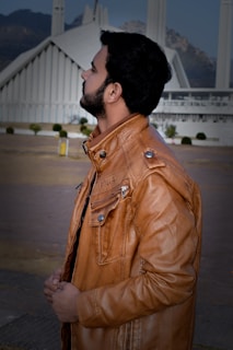 A man with a beard stands outdoors, wearing a light brown leather jacket with the initials 'D&G' on it. Behind him is an architectural structure that resembles a modern mosque with tall, white spires and mountains in the background.