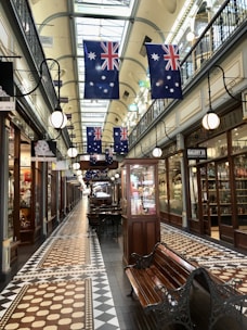An indoor shopping arcade with a vintage and elegant design. The ceiling is high and adorned with skylights that allow natural light to illuminate the space. Numerous Australian flags hang from the ceiling, adding a patriotic touch to the corridor. The floor features intricate tile patterns in shades of brown and beige. On both sides of the walkway, there are shops with glass windows displaying various merchandise. Benches are placed along the central walkway, offering a place for shoppers to rest. The ambiance is complemented by classic street-style lamps attached to the walls.