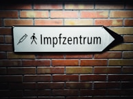 A white arrow sign mounted on a brick wall points to the right. The sign features a syringe icon and a pedestrian symbol, with the text 'Impfzentrum' indicating a vaccination center.
