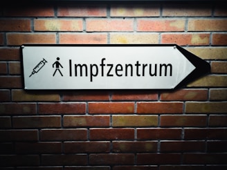 A white arrow sign mounted on a brick wall points to the right. The sign features a syringe icon and a pedestrian symbol, with the text 'Impfzentrum' indicating a vaccination center.