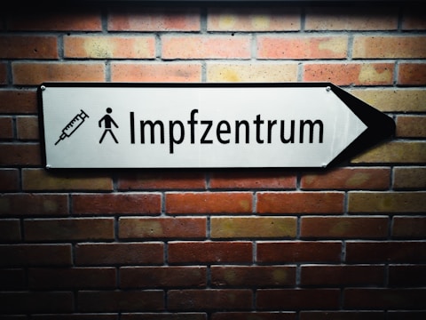 A white arrow sign mounted on a brick wall points to the right. The sign features a syringe icon and a pedestrian symbol, with the text 'Impfzentrum' indicating a vaccination center.
