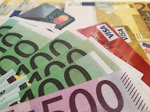 A close-up view of various Euro banknotes and credit cards scattered together. The banknotes have denominations of 500 and 100 Euros, while the visible credit cards include both Mastercard and Visa. The composition suggests a theme related to finance and currency.