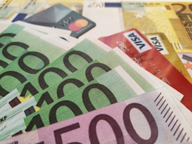 A close-up view of various Euro banknotes and credit cards scattered together. The banknotes have denominations of 500 and 100 Euros, while the visible credit cards include both Mastercard and Visa. The composition suggests a theme related to finance and currency.