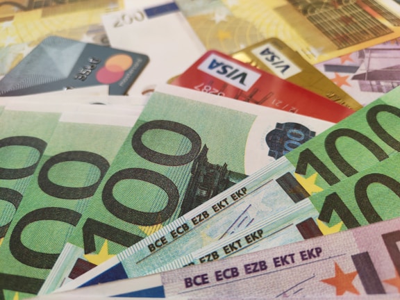A close-up of various euro banknotes and credit cards arranged together. The focus is on the 100 euro bills, with multiple denominations of euro bills and several credit cards, including VISA and Mastercard, scattered in the background.