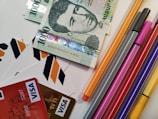 A selection of various colored markers and pens is arranged next to a stack of ten thousand Hungarian forint banknotes. Several credit cards with Visa logos are visible, along with envelopes featuring orange, black, and white chevron patterns.