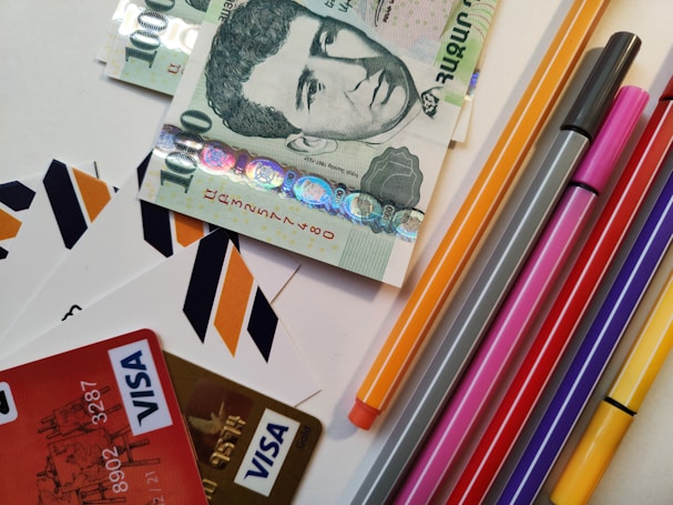 A selection of various colored markers and pens is arranged next to a stack of ten thousand Hungarian forint banknotes. Several credit cards with Visa logos are visible, along with envelopes featuring orange, black, and white chevron patterns.