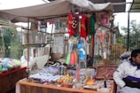 An outdoor market stall is filled with a variety of colorful trinkets and accessories. The stall has strings of beads and necklaces hanging from the top, and numerous small items such as bracelets, keychains, and decorative pieces are displayed on tables. The vendor is sitting on the right side, dressed in a white and purple jacket. The background features a fence and foliage, indicating an open-air setting.