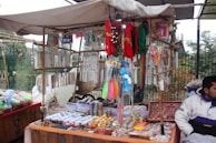 An outdoor market stall is filled with a variety of colorful trinkets and accessories. The stall has strings of beads and necklaces hanging from the top, and numerous small items such as bracelets, keychains, and decorative pieces are displayed on tables. The vendor is sitting on the right side, dressed in a white and purple jacket. The background features a fence and foliage, indicating an open-air setting.