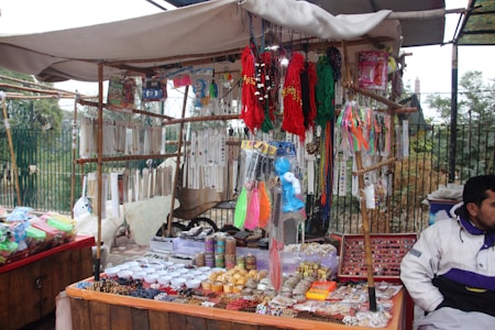 An outdoor market stall is filled with a variety of colorful trinkets and accessories. The stall has strings of beads and necklaces hanging from the top, and numerous small items such as bracelets, keychains, and decorative pieces are displayed on tables. The vendor is sitting on the right side, dressed in a white and purple jacket. The background features a fence and foliage, indicating an open-air setting.