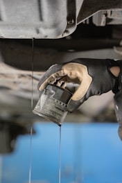 A gloved hand is holding an oil filter as oil pours out into a stream. The background includes parts of a vehicle's undercarriage, likely indicating an oil change process.