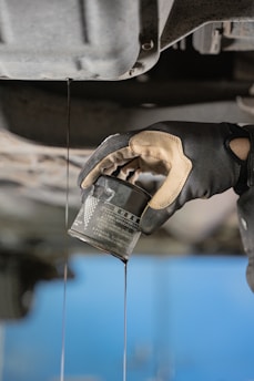 A gloved hand is holding an oil filter as oil pours out into a stream. The background includes parts of a vehicle's undercarriage, likely indicating an oil change process.